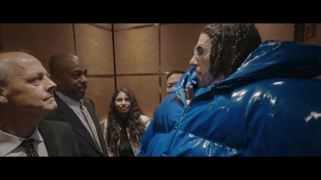 Ali G in een blauwe pufferjas in een lift vol mannen in pak, uit de trailer van Ali G: Who Iz I?