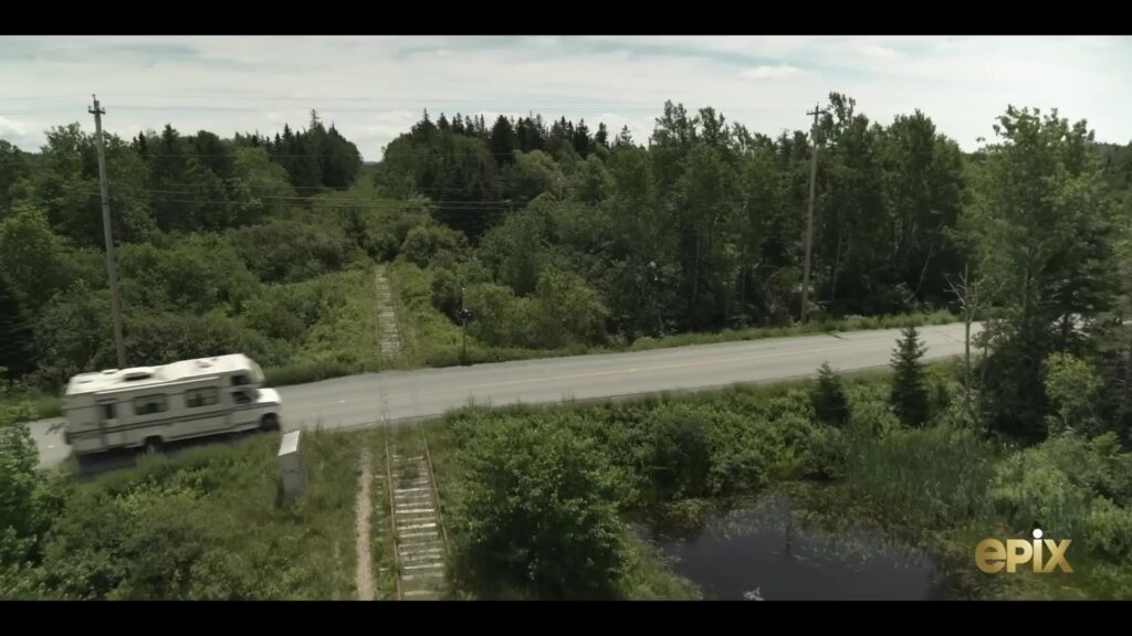 Een camper rijdt over een landweg tussen de bossen in de horrorserie From