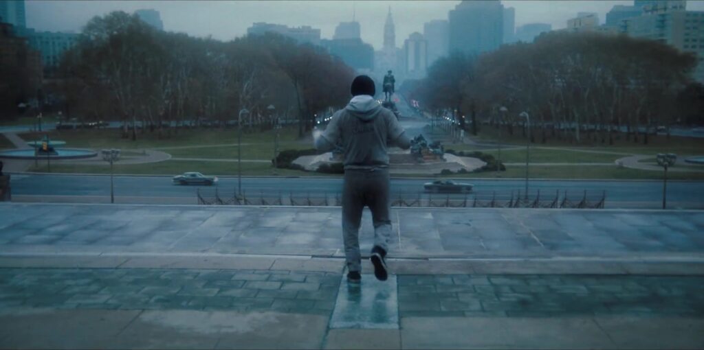 Een jonge bokser in grijs trainingspak rent de trappen op in Philadelphia in de trailer van I Play Rocky