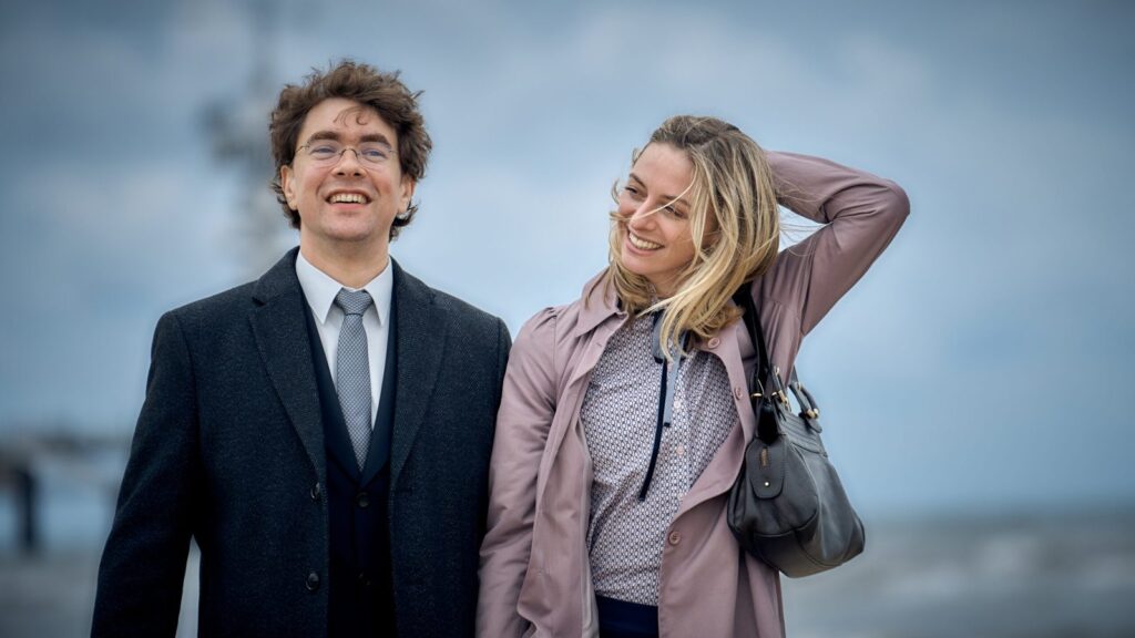 Een jonge man in pak en een lachende vrouw op een winderige pier in een eerste still uit Mabel & Margarita